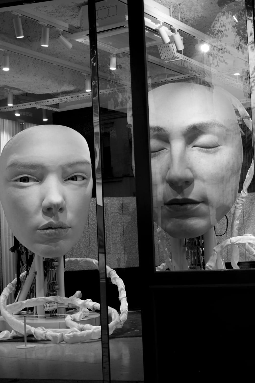 Fotografia de Paulo Ricardo Santos: dois rostos escultóricos atrás de uma vitrine, um deles refletido no vidro, em preto e branco