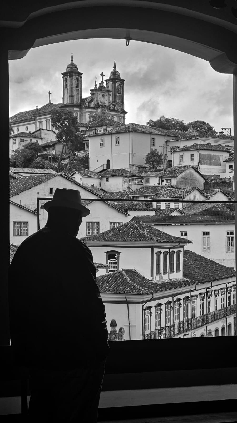 Fotografia de Paulo Ricardo Santos: o vulto de uma pessoa de chapéu diante de telhados e torres de igreja, em preto e branco
